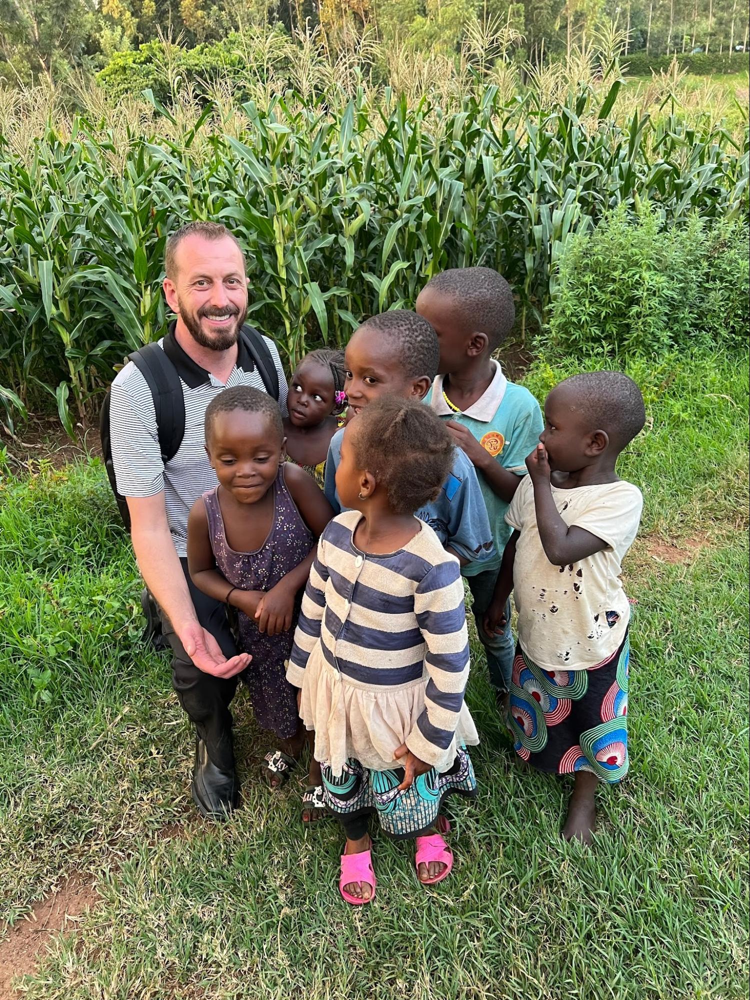 Team member kneeling with a group of children beside a maize field