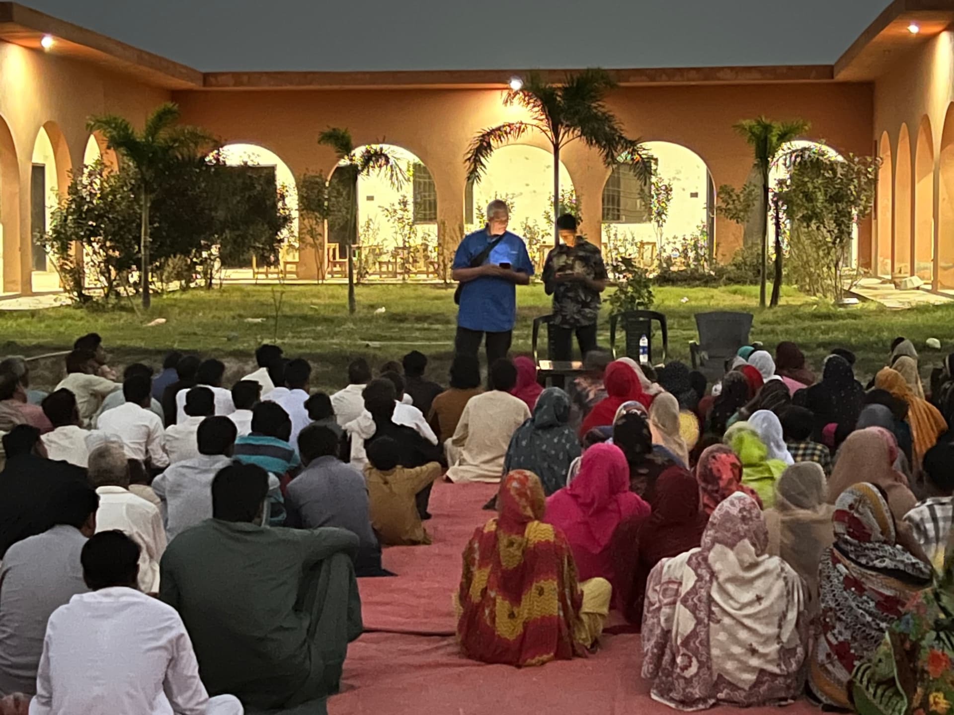 An evening outdoor outreach gathering with a speaker addressing a seated crowd in Pakistan